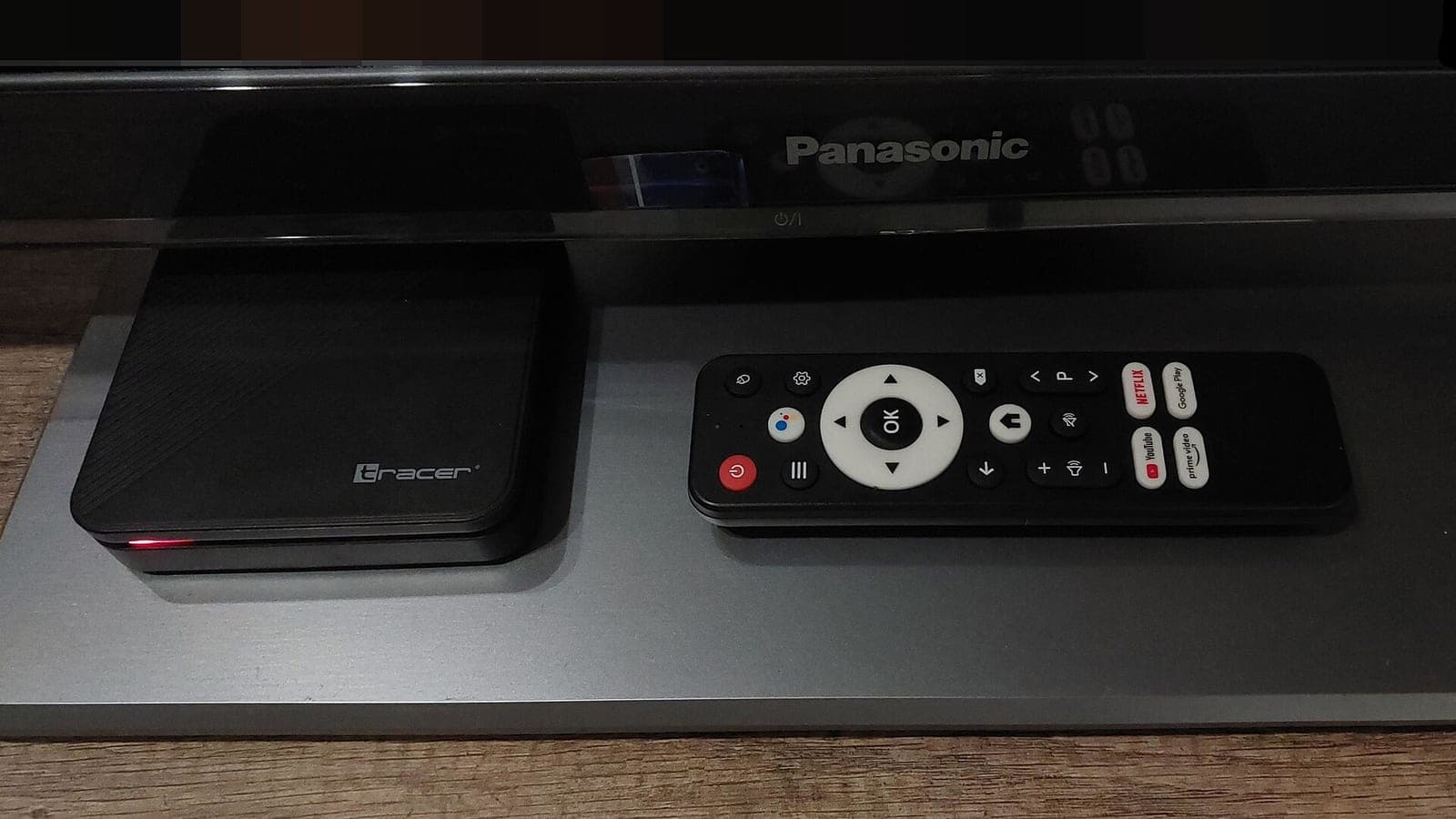 A small black Android TV set-top box and its remote control on a stand under a television