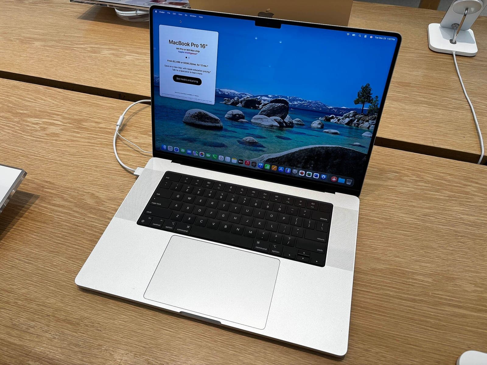 A silver 16-inch MacBook Pro with an M4 Pro chip open on a wooden table in an Apple Store, showing the macOS desktop