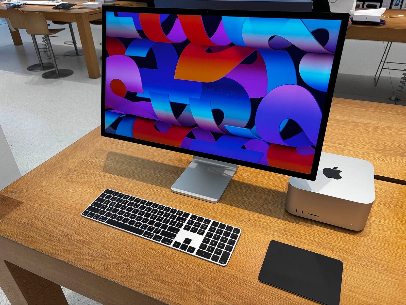 A silver Mac Studio desktop computer next to an Apple Studio Display with a colorful wallpaper, a black and white keyboard and a black trackpad on a wooden table in an Apple Store