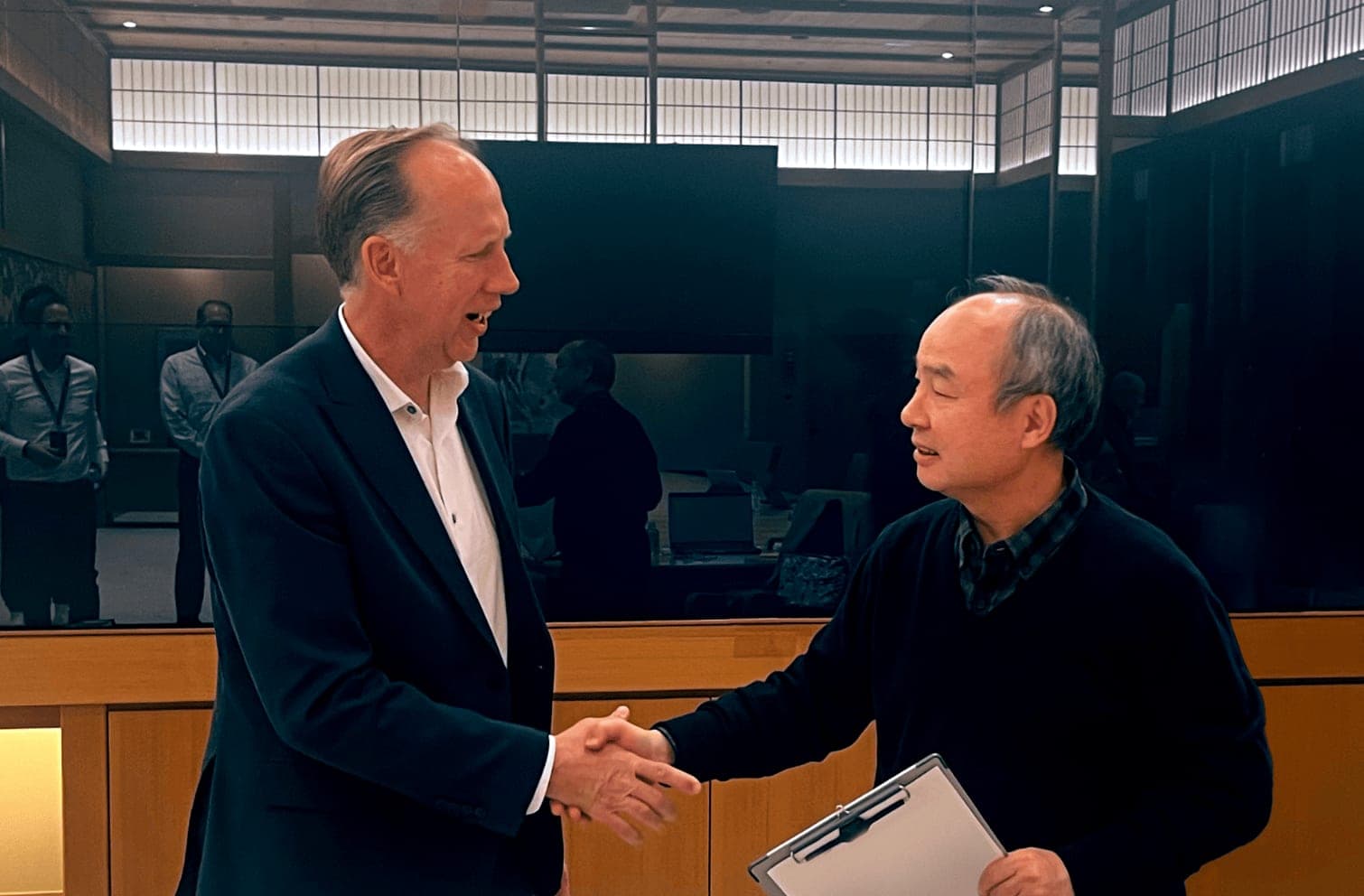 Two men in business attire shaking hands in a meeting room, one holding a clipboard