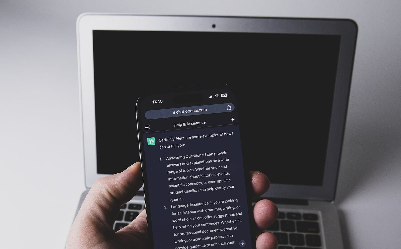 A hand holding a smartphone showing a ChatGPT conversation, with an open laptop in the background