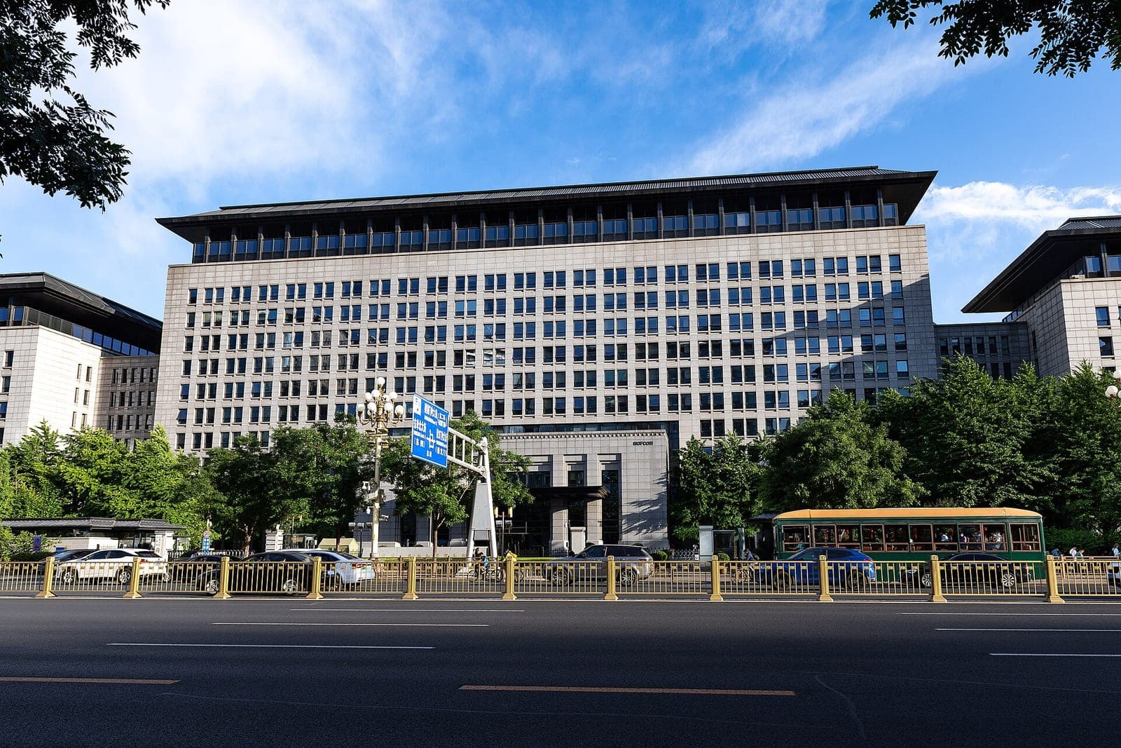 The large stone headquarters of China's Ministry of Commerce on a wide avenue in Beijing, with trees, a gold-colored fence and a green bus in front under a blue sky