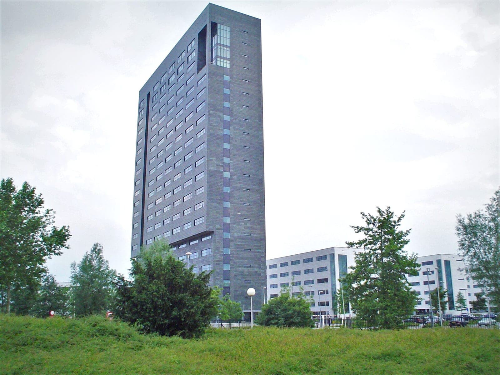 ASML's dark, slab-shaped office tower in Veldhoven, the Netherlands, rising behind trees and a grassy slope, with lower white office buildings beside it