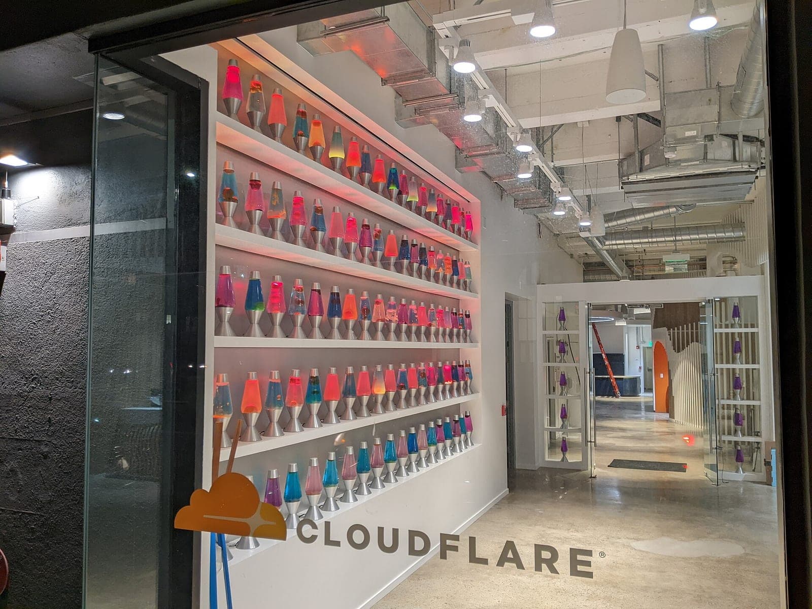 A wall of colorful lava lamps on white shelves behind a glass door with the Cloudflare name at a Cloudflare office