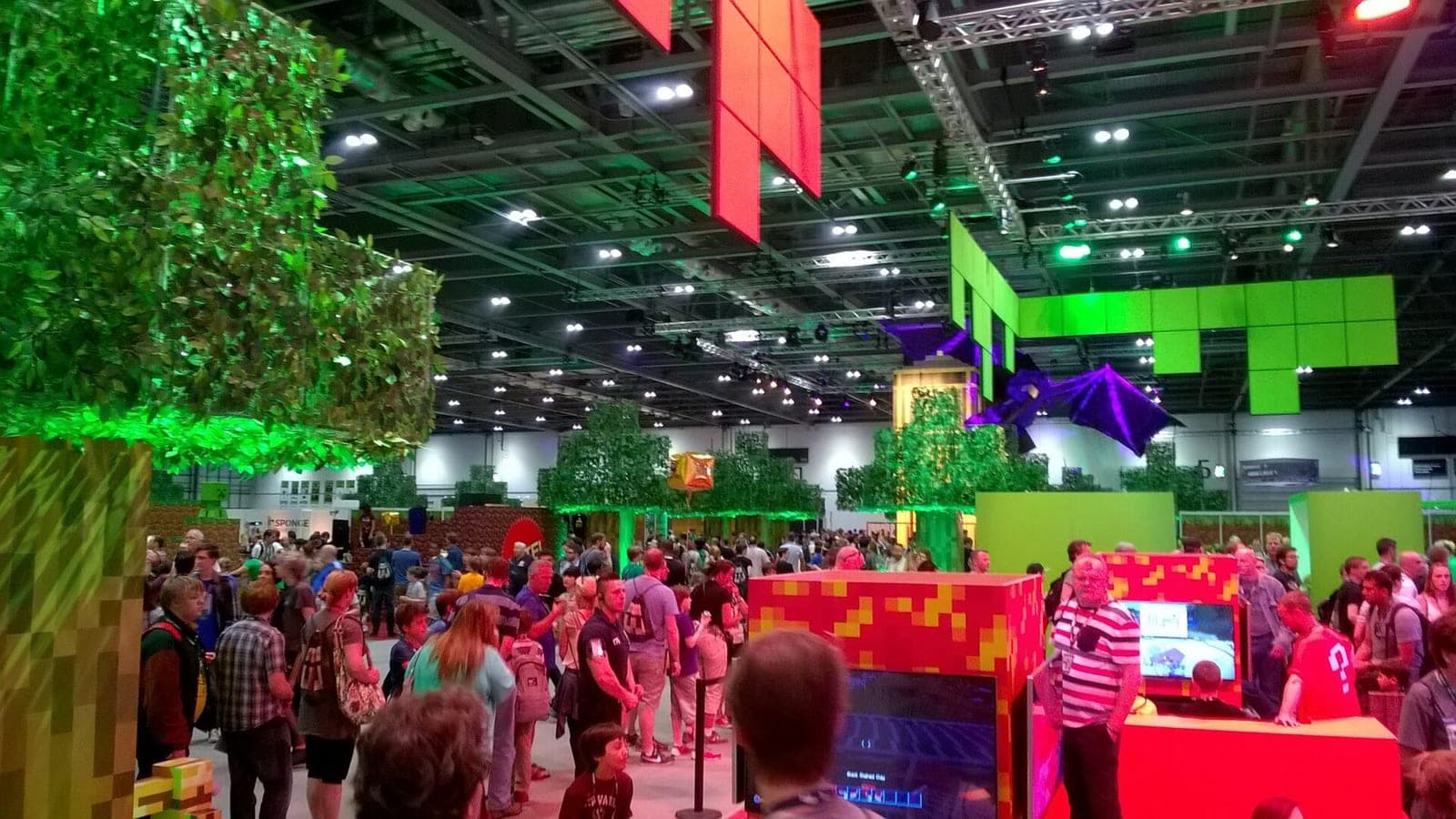 Crowds of fans in the MineCon 2015 expo hall, with giant Minecraft trees, blocky booths and an Ender Dragon model hanging from the ceiling