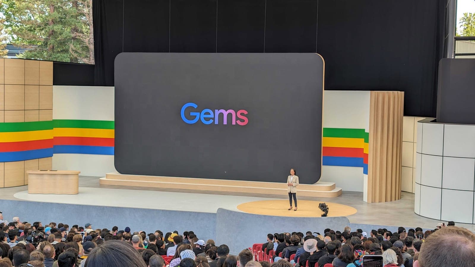 A presenter on stage at a Google event in front of a large screen displaying the word 'Gems' in colorful gradient text, with an audience seated in front.