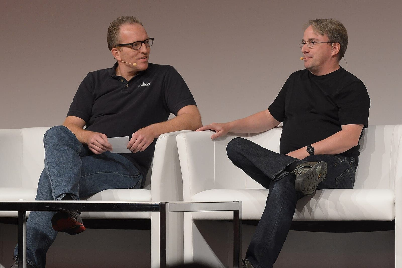 Linus Torvalds, right, in a black T-shirt, sitting on a white armchair on stage at LinuxCon Europe during an onstage conversation