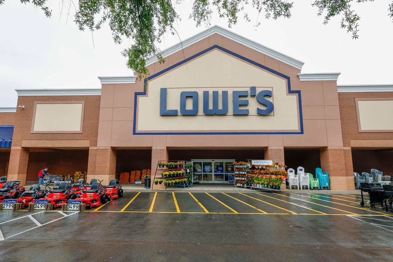 The exterior of a Lowe's home improvement store with its blue and tan sign, parking lot, and outdoor garden display.