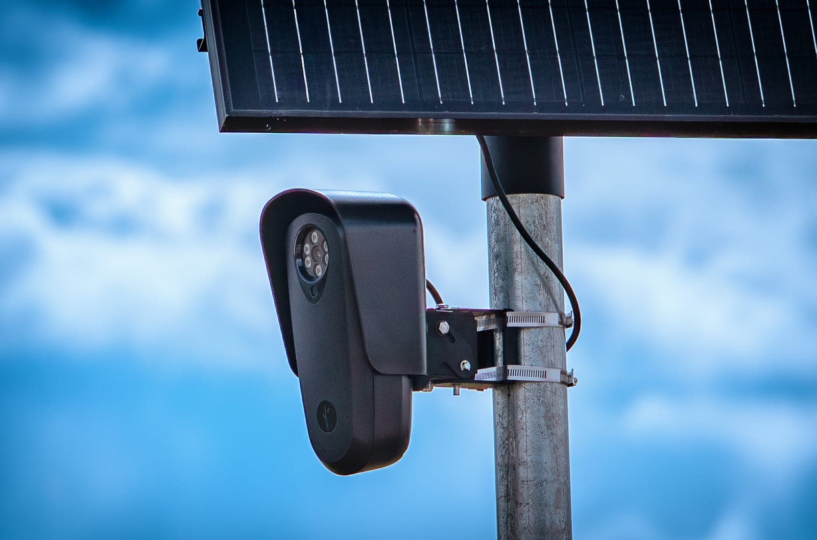 A black Flock Safety license plate reader camera mounted on a metal pole under a solar panel, against a blue sky