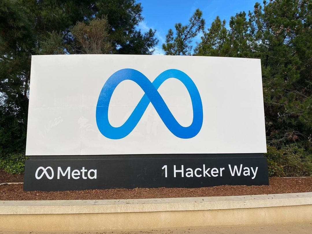 A white sign with Meta's blue infinity logo and the address 1 Hacker Way outside the company's headquarters in Menlo Park, surrounded by pine trees.