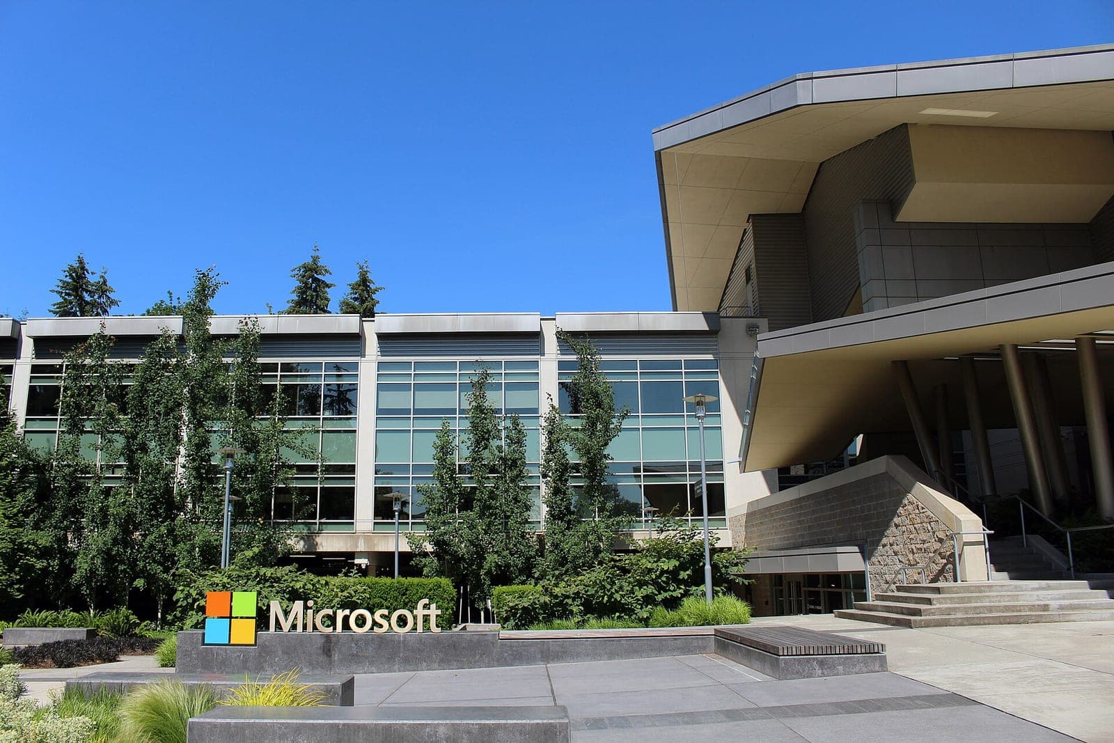 A Microsoft sign with the four-color logo in front of a glass office building and a modern entrance on the company's Redmond campus on a sunny day