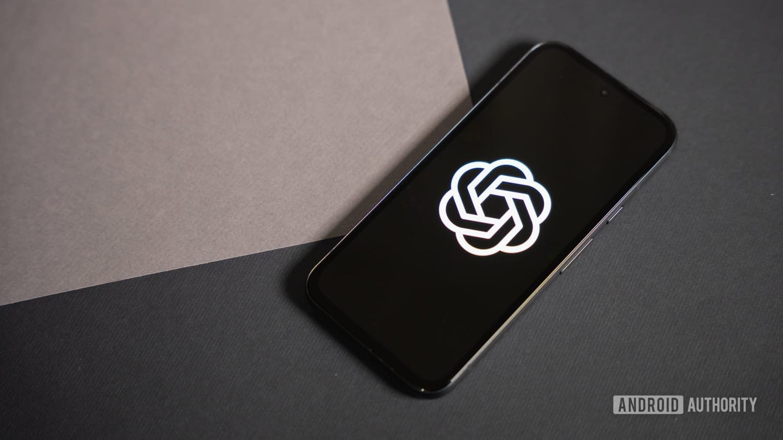 A smartphone lying on a two-tone paper background displaying the glowing white OpenAI logo on its screen.