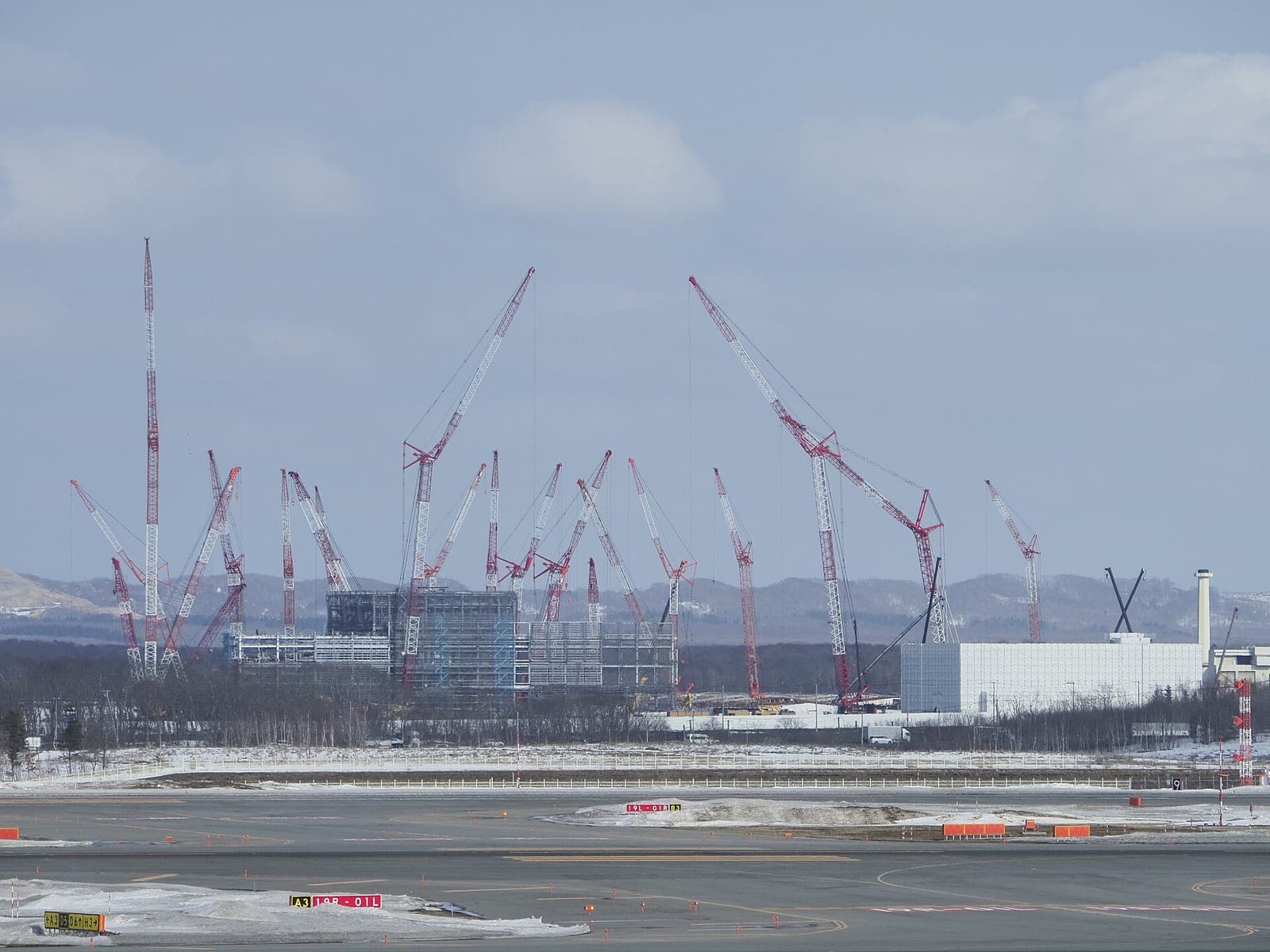 Dozens of red and white construction cranes over the Rapidus chip factory being built in Japan, seen across a snowy airport apron