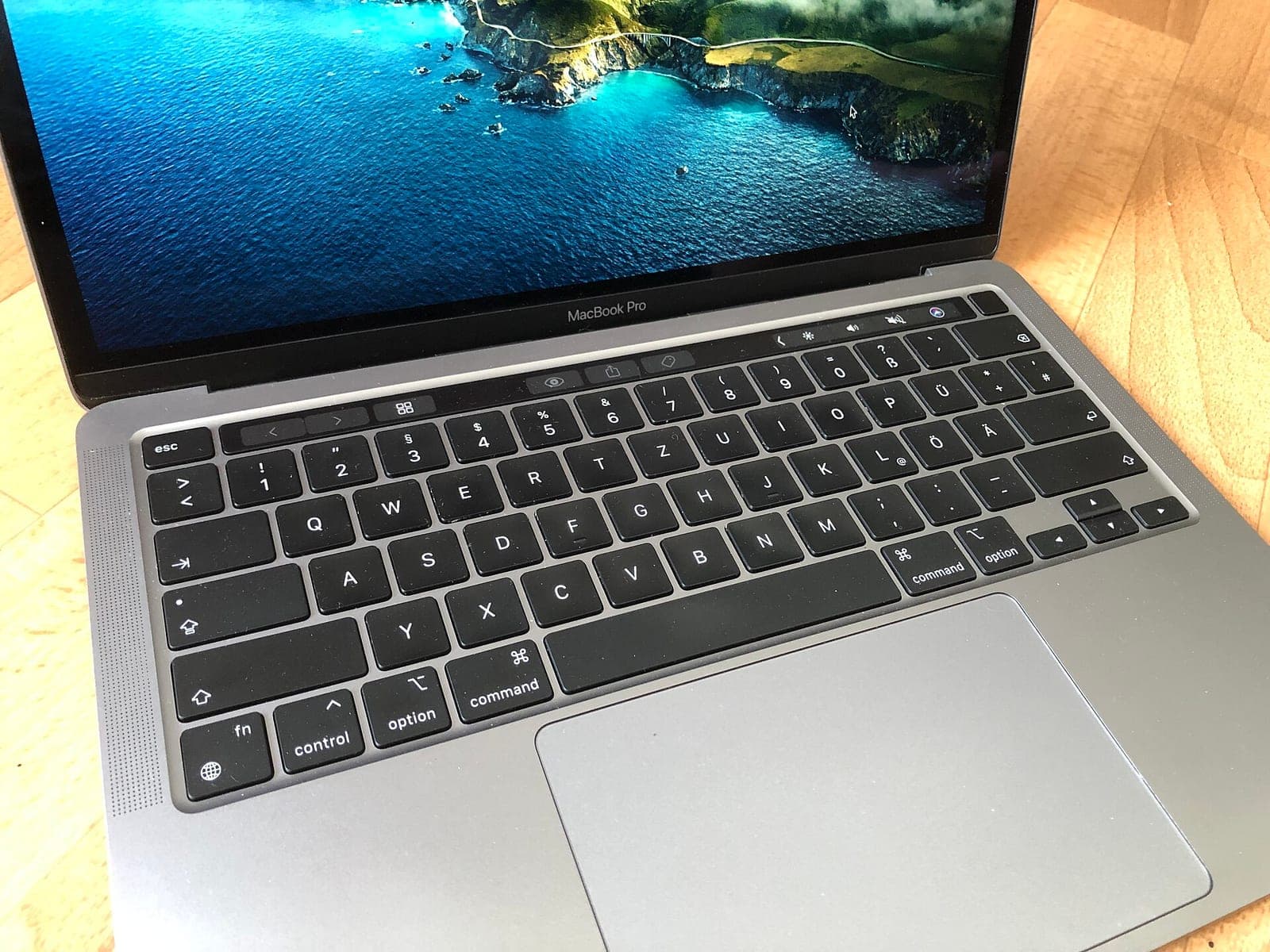 A 13-inch MacBook Pro with Apple silicon and a Touch Bar, open on a wooden table with a coastal wallpaper on screen