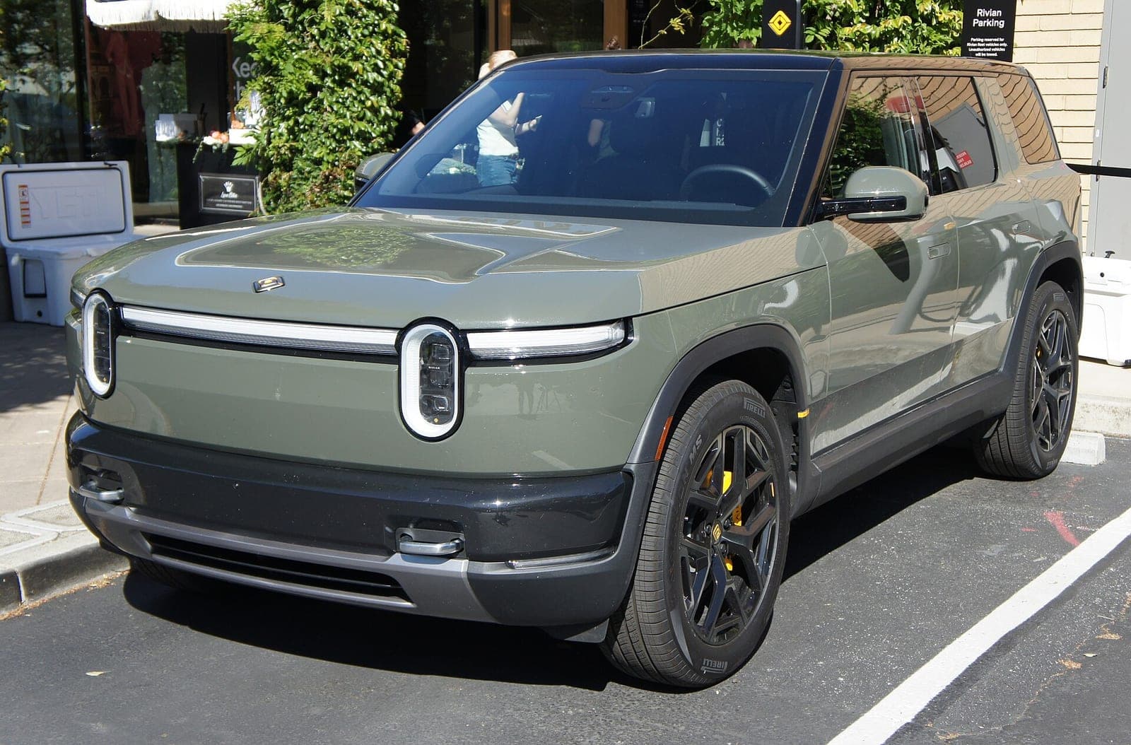 A green Rivian R2 electric SUV parked at the curb outside a Rivian location, with its oval headlights and full-width light bar