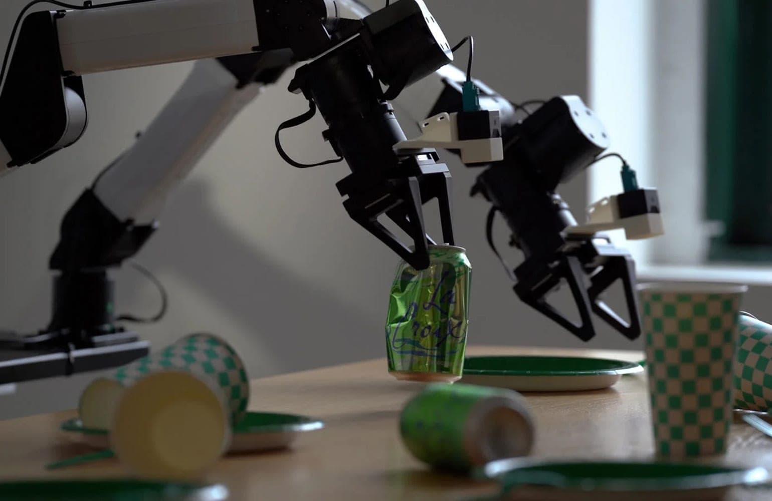 Two black and white robotic arms on a table, one gripper lifting a green La Croix can among checkered paper cups and plates