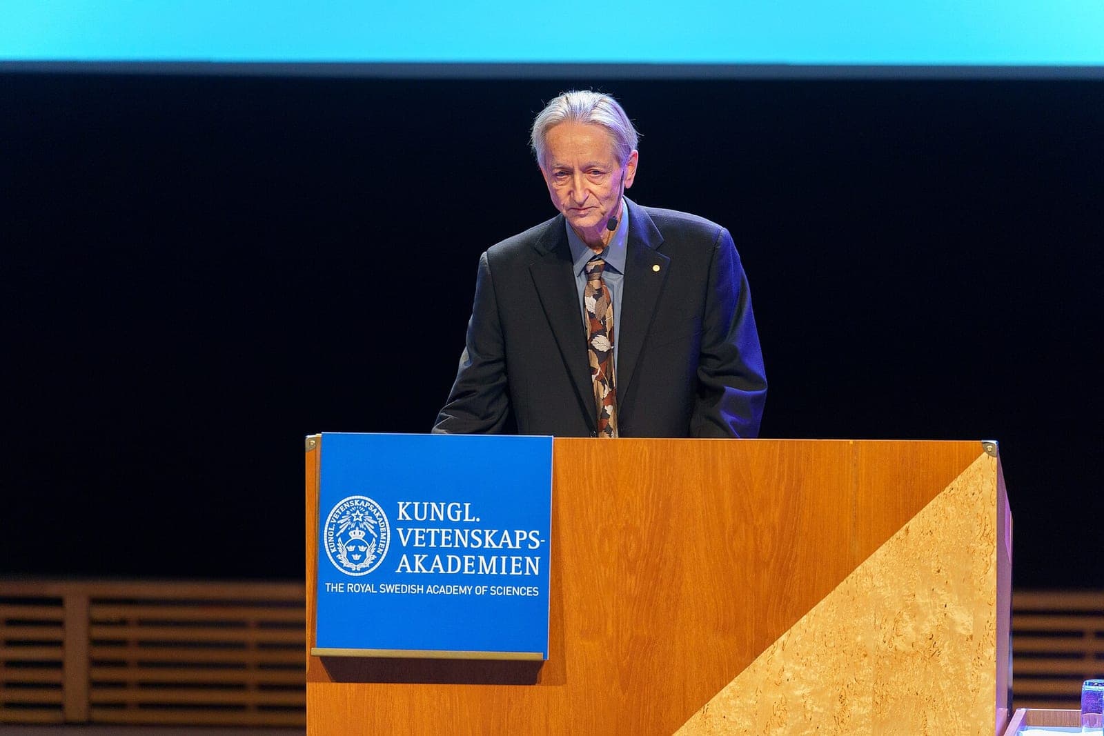 Geoffrey Hinton, in a dark suit and patterned tie, speaks at a wooden podium marked with the Royal Swedish Academy of Sciences sign during his 2024 Nobel lecture