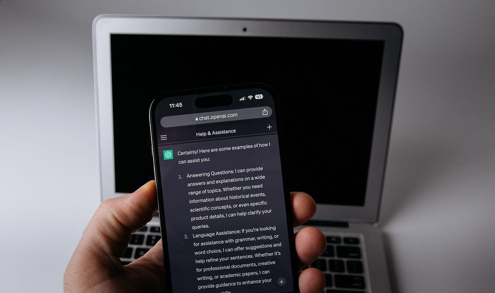 A hand holds a smartphone showing ChatGPT in a web browser, with an open laptop in the background