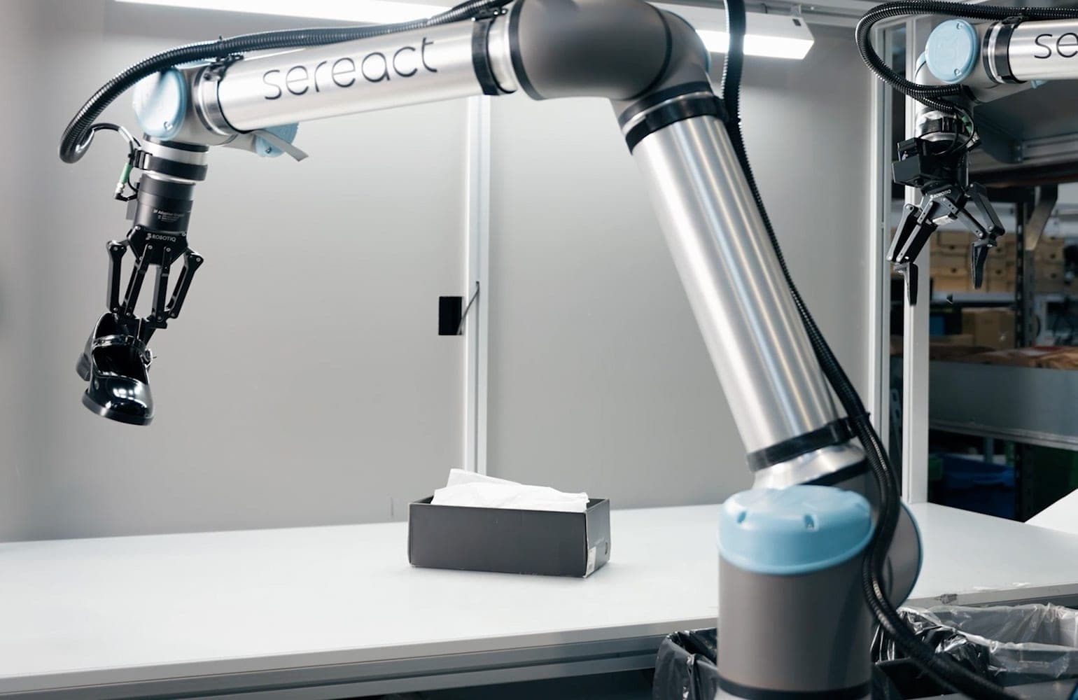 A Sereact-branded Universal Robots arm holds a black leather shoe in a gripper above a table with an open box, with a second arm and warehouse shelves behind it