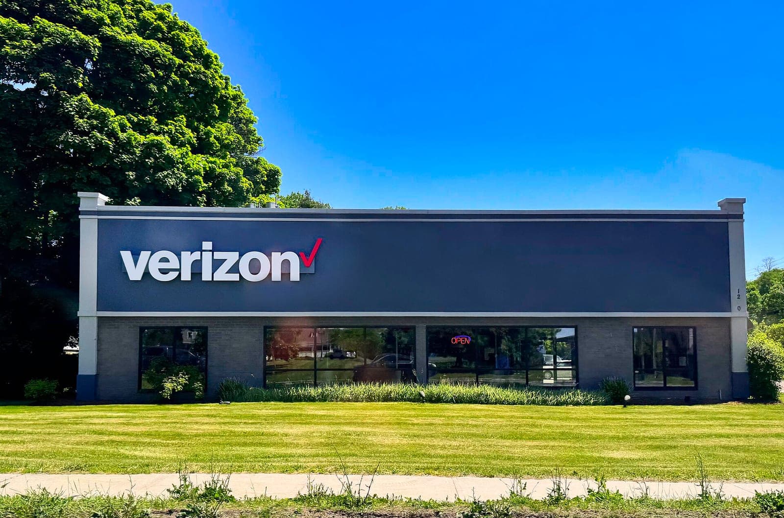 The front of a Verizon retail store with a large white Verizon sign, big windows and an open sign, behind a mowed lawn under a clear blue sky