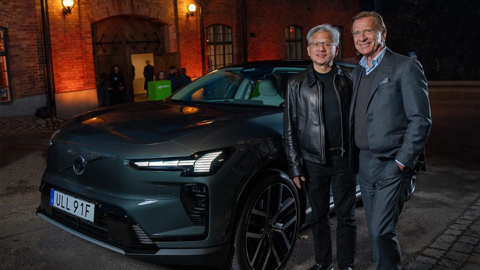 Nvidia CEO Jensen Huang in a black leather jacket and Volvo Cars CEO Hakan Samuelsson in a grey suit standing beside a dark green Volvo electric SUV outside a brick building at night