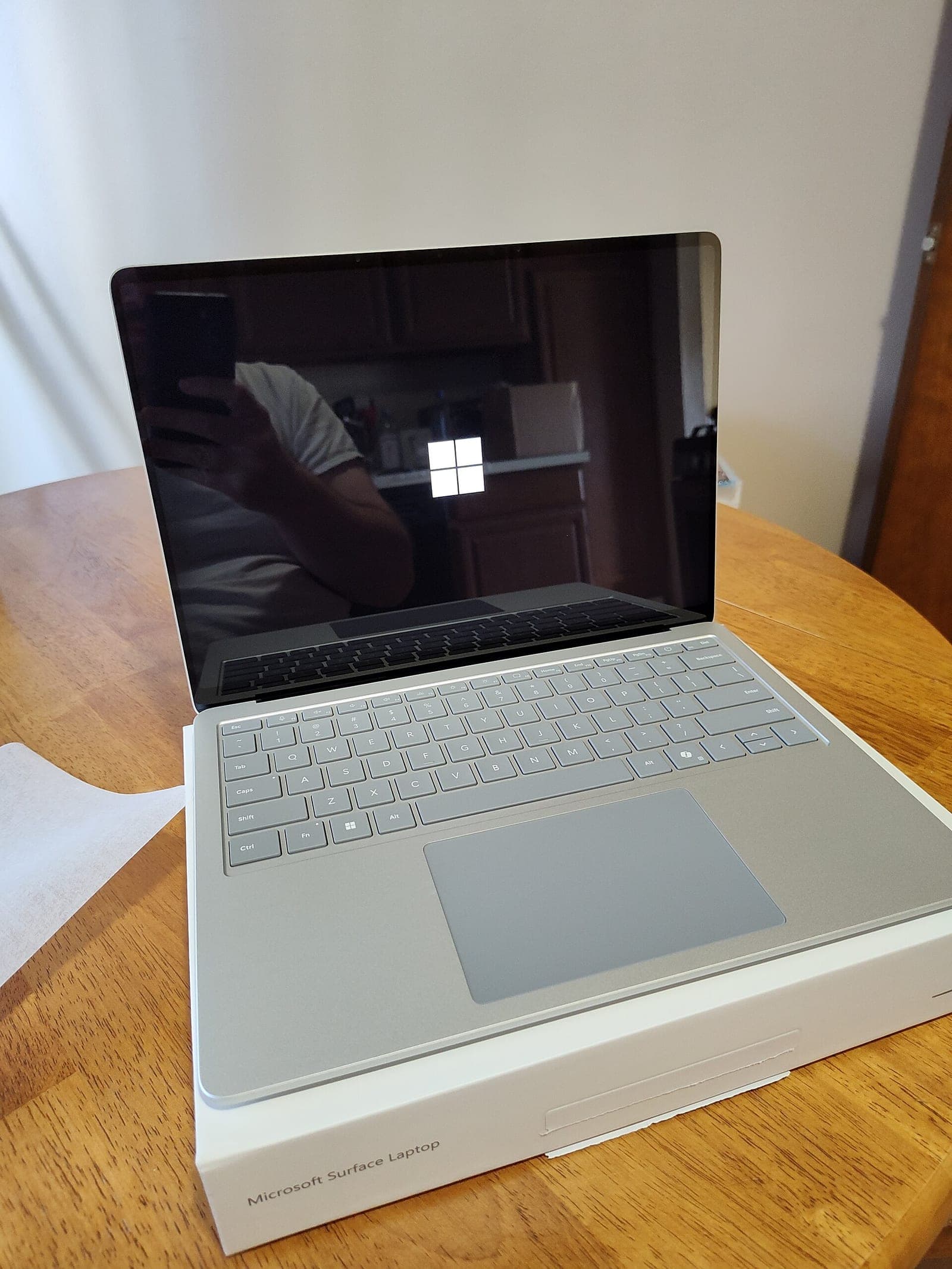A silver Microsoft Surface Laptop sitting open on its box on a wooden table, with the Windows logo on its screen during startup