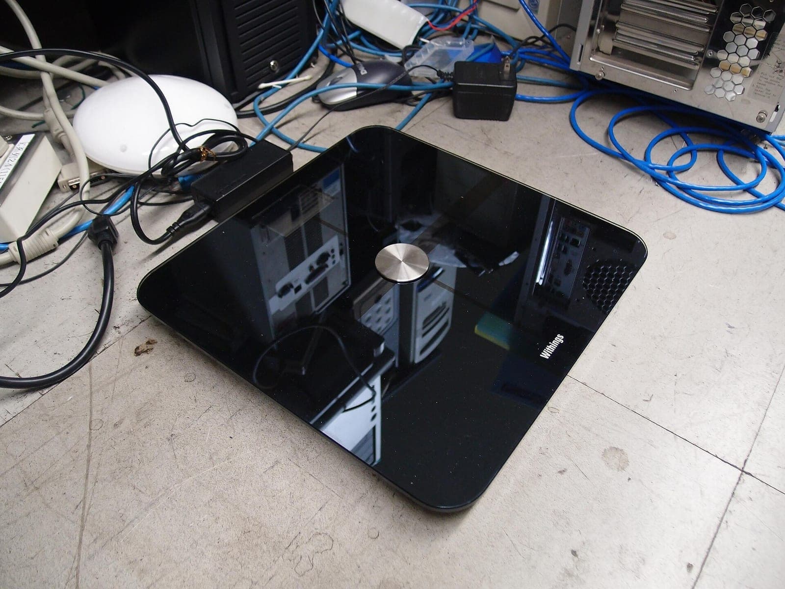 A black Withings Wi-Fi body scale with a round metal button in the middle, sitting on a concrete floor surrounded by computer cables and power adapters