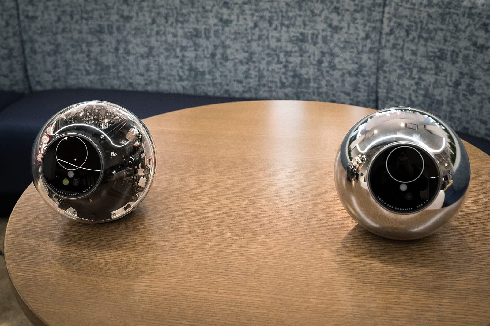 Two of World's spherical Orb devices, one with a clear shell showing its circuit boards and one in polished chrome, sitting on a round wooden table