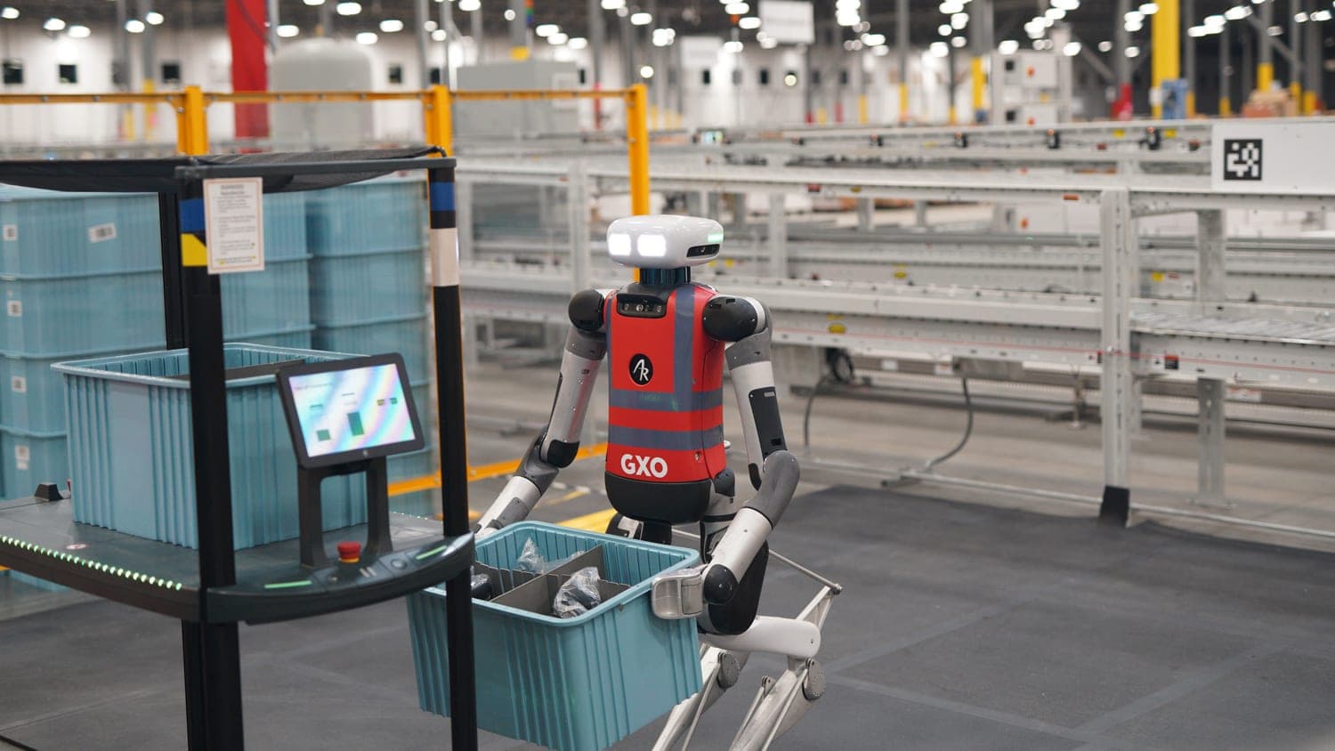 An Agility Robotics Digit humanoid, the earlier model with backward-bending legs, wearing a red GXO vest and carrying a blue tote past a workstation in a large warehouse