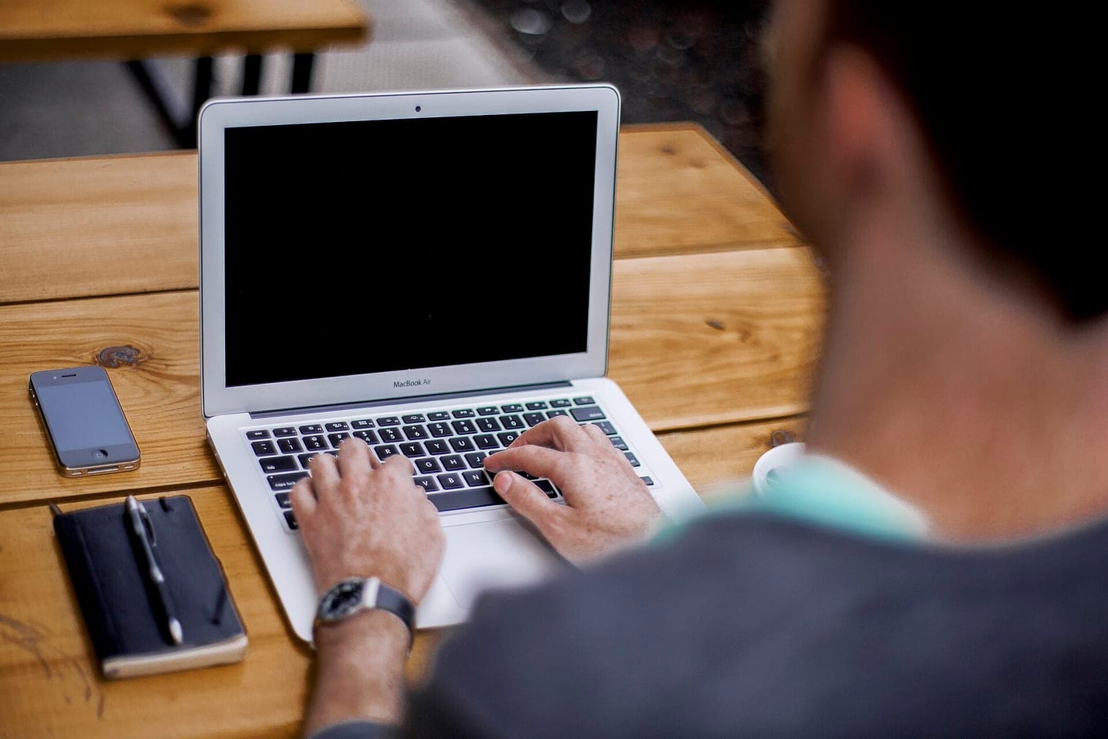 A person's hands typing on a laptop keyboard at an outdoor wooden table, with a phone and notebook nearby