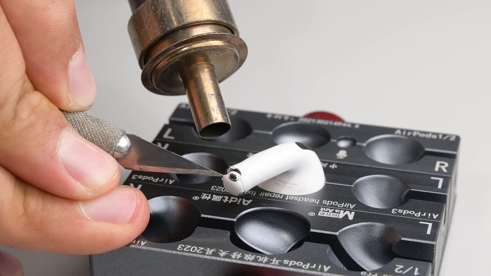 A technician using a heat tool and tweezers to disassemble a white AirPod on a repair jig