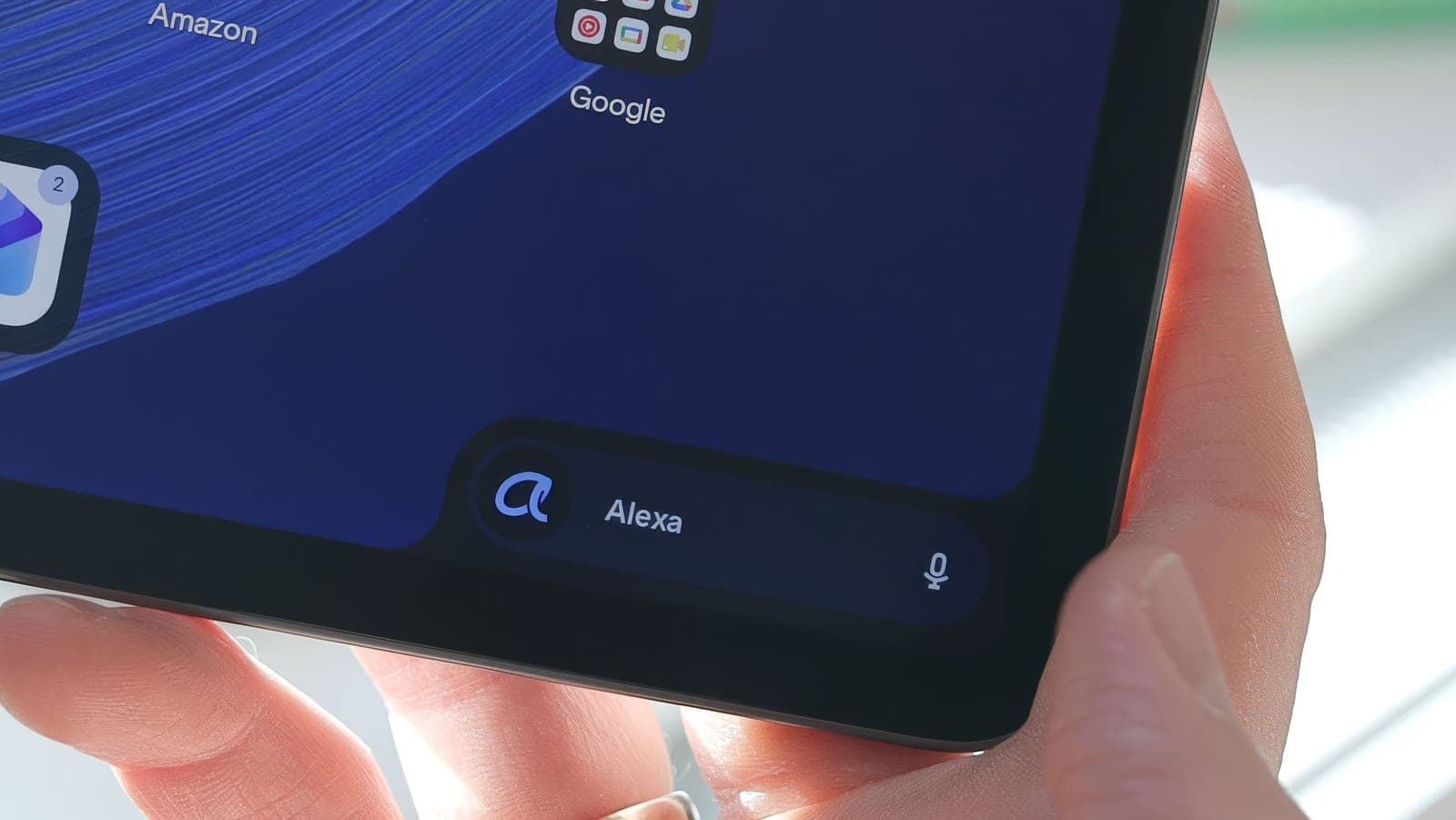 A hand holding the Amazon Alexa Tablet 12 Pro, close up on the lower corner of the home screen with the Ask Alexa bar and a microphone icon