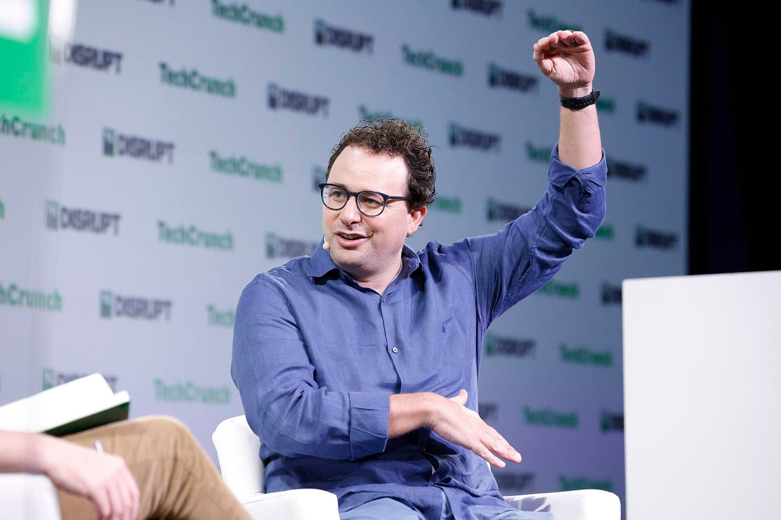 Anthropic CEO Dario Amodei, in glasses and a blue shirt, raises one hand while speaking on stage at TechCrunch Disrupt 2023