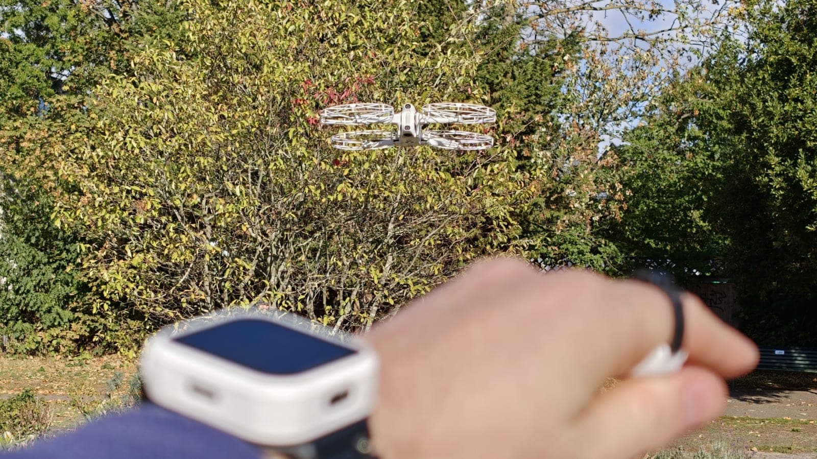 A blurred wrist wearing the white Antigravity watch remote and a ring remote, with the A1 drone hovering in front of autumn trees
