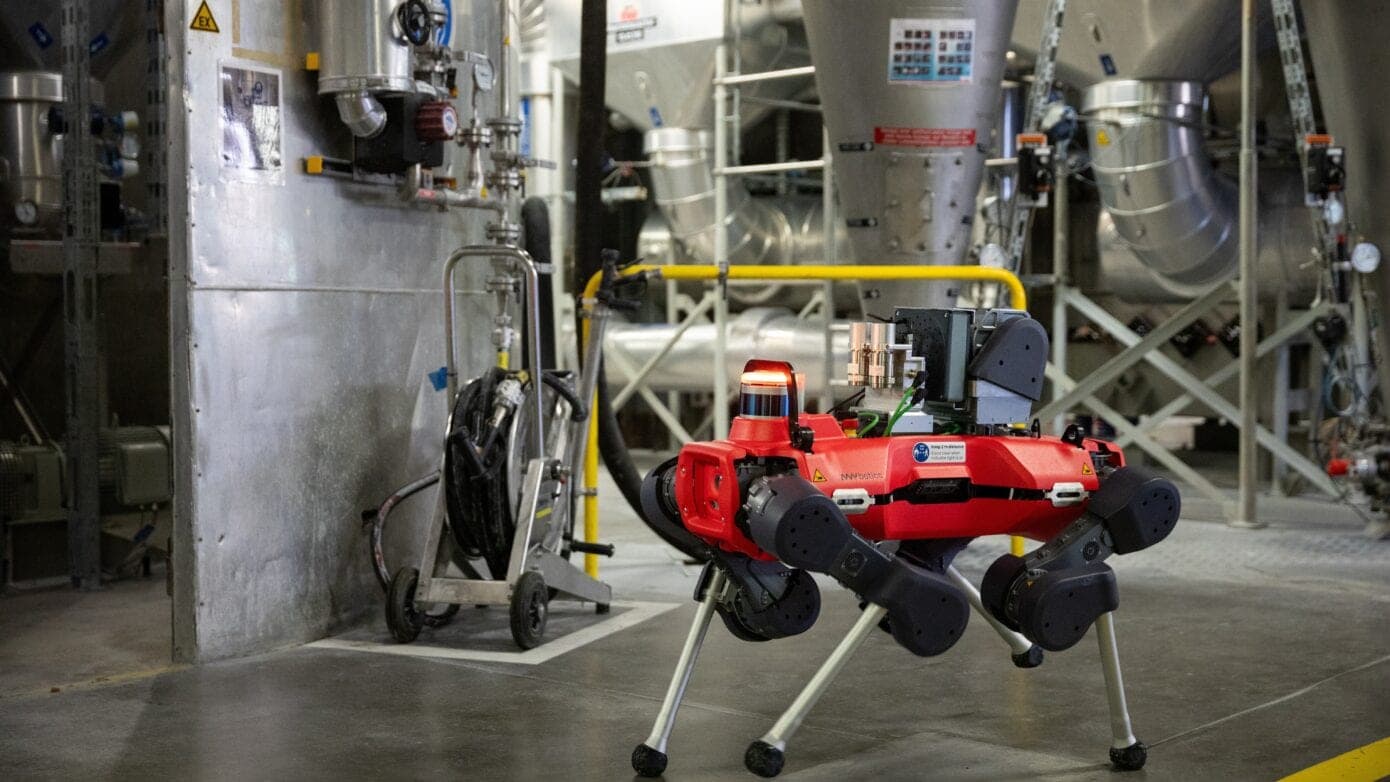 A red four-legged ANYmal inspection robot walking through an industrial facility with large metal pipes and tanks