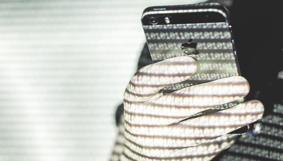 A gloved hand holding an iPhone with cascading binary code projected across the phone and hand in a darkened room.