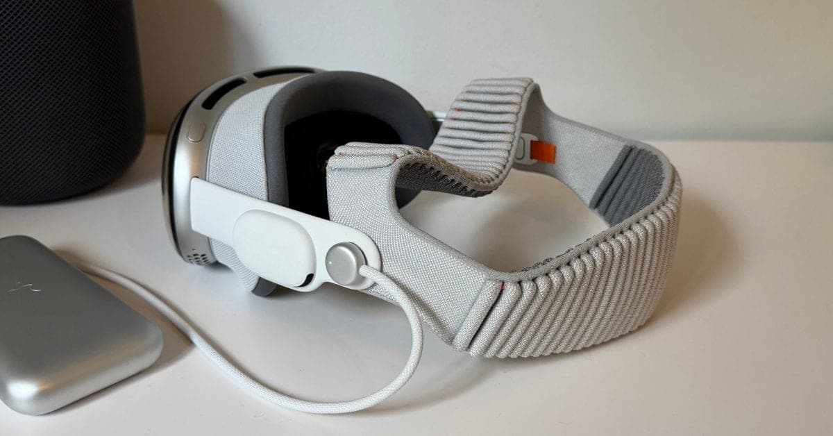 A white Apple Vision Pro headset with its fabric strap, sitting on a white table next to a HomePod and a battery pack.