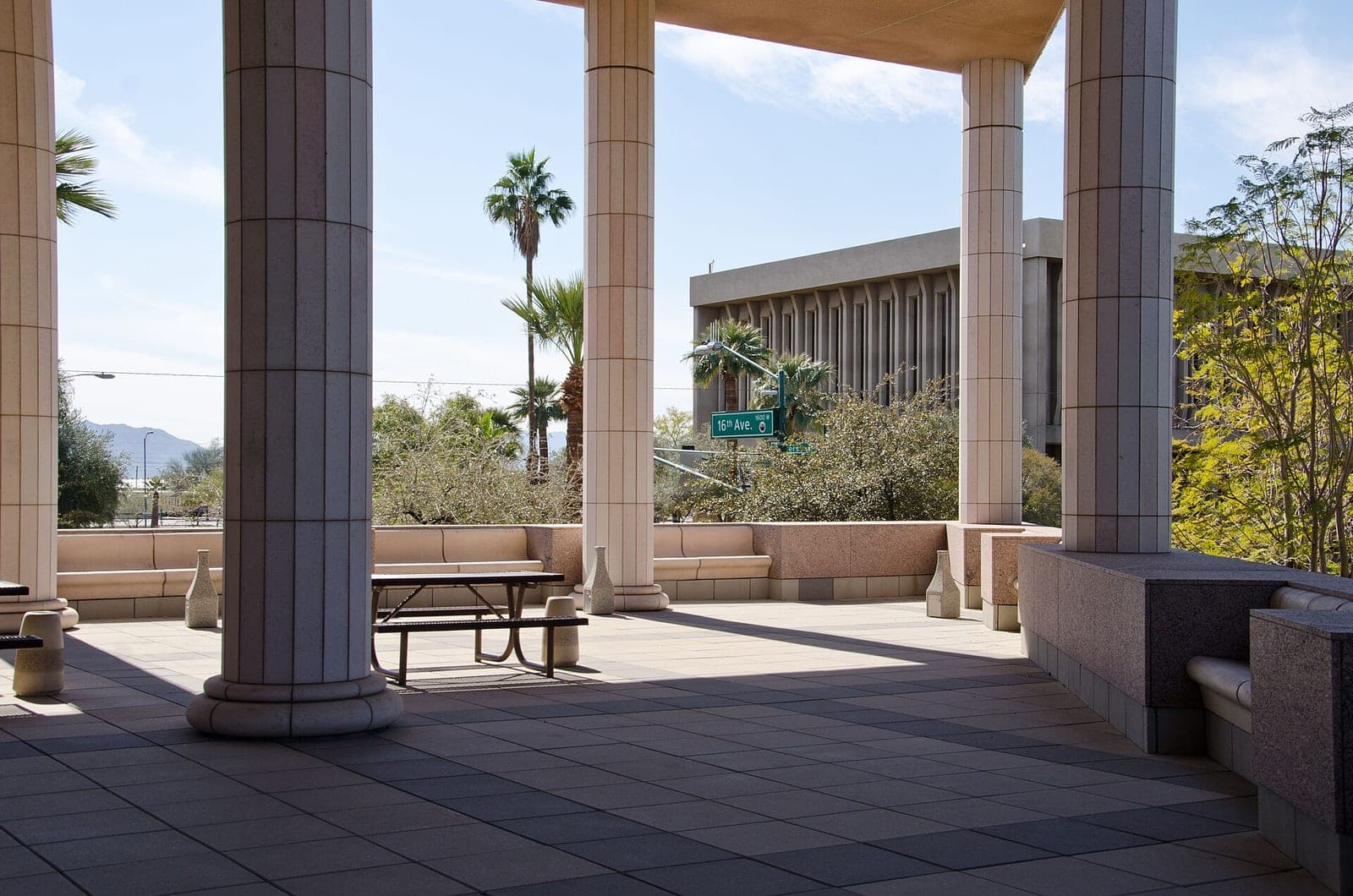 The shaded colonnade outside the Arizona Supreme Court building in Phoenix, with stone columns, a picnic table and a 16th Ave. street sign