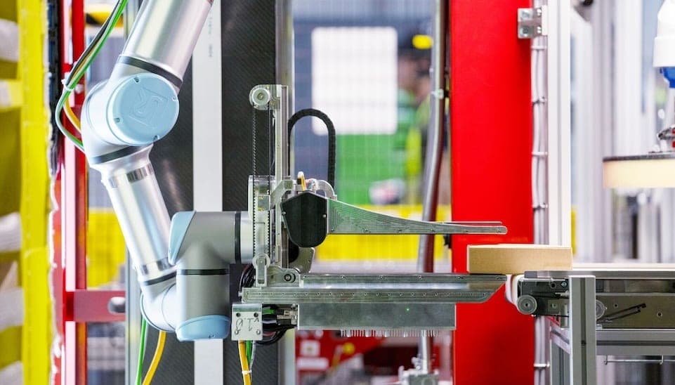 A collaborative robot arm with a custom flat gripper sliding a cardboard box onto a conveyor inside an automated warehouse cell.
