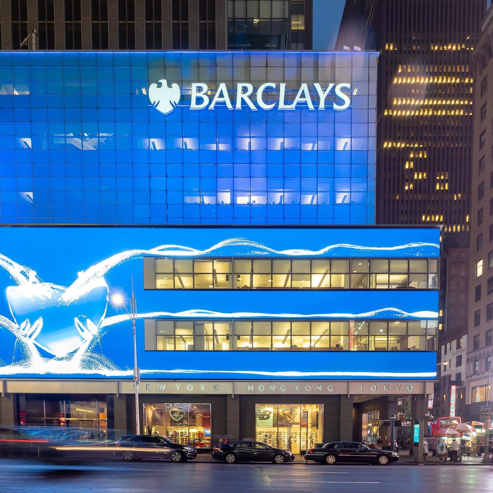 The illuminated Barclays headquarters building at night in Midtown Manhattan, with the Barclays logo lit up above large glass windows