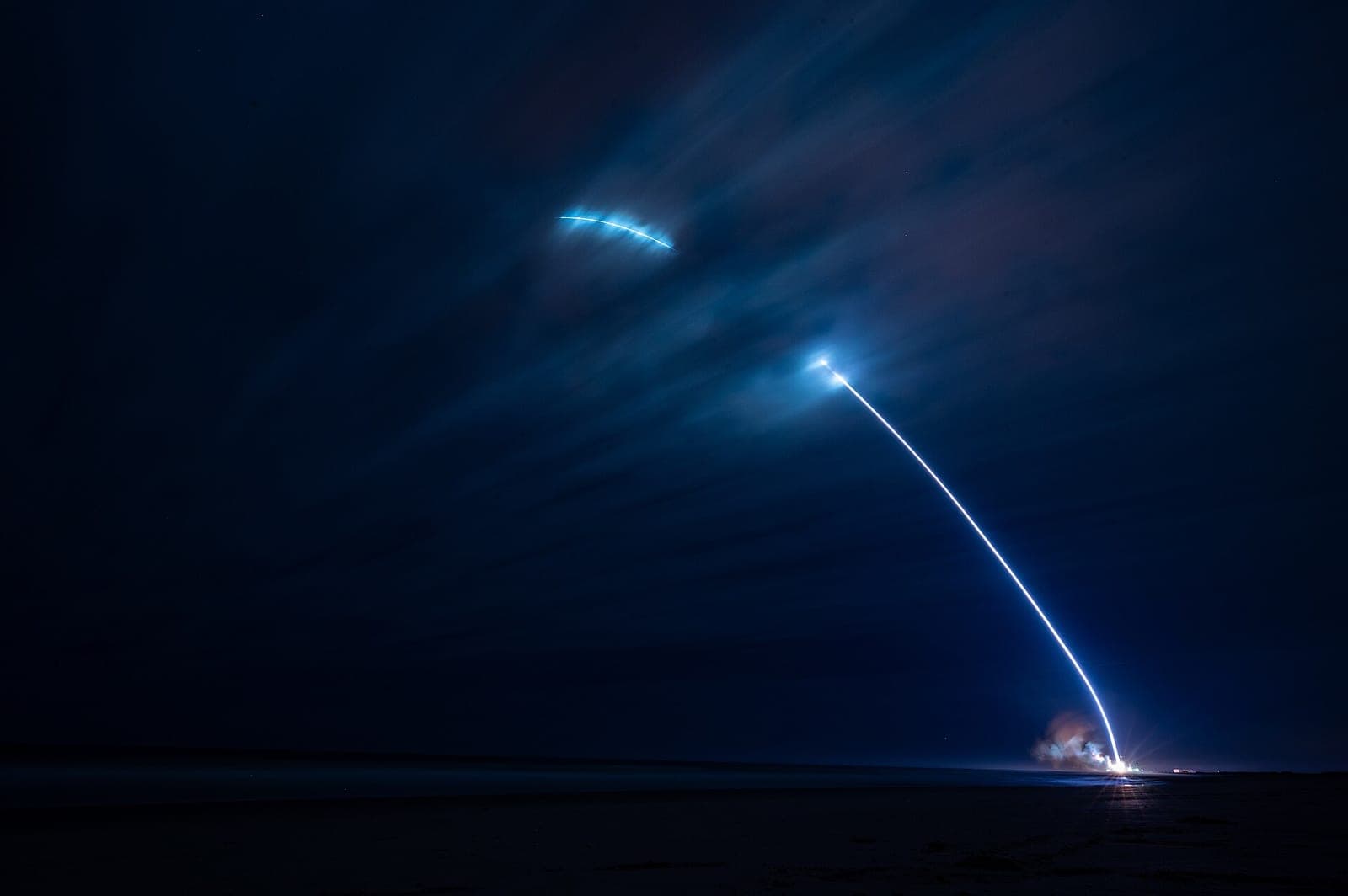 Long exposure photo of a New Glenn rocket launching at night from Space Launch Complex 36, leaving a bright curved trail across a dark blue sky