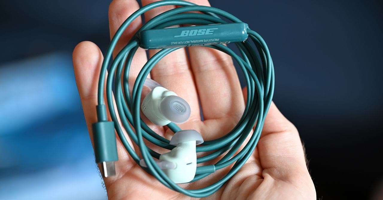 A hand holding the coiled teal cable of Bose's new wired earbuds, showing the Bose logo and the white earbud tips.