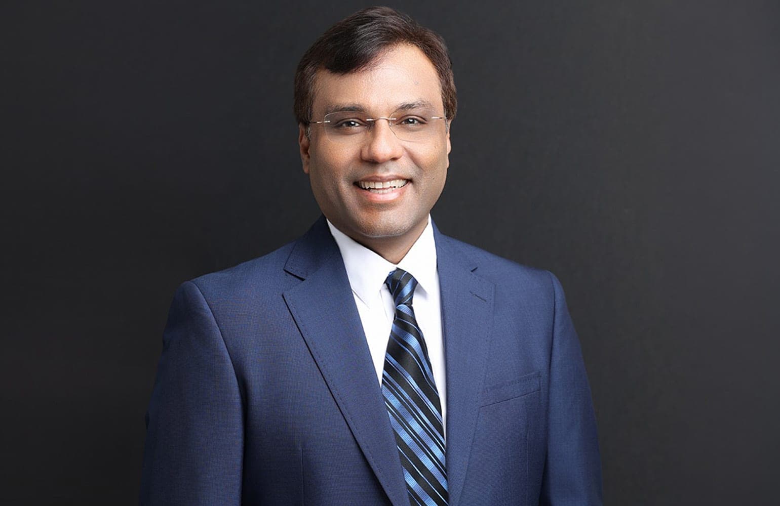 Portrait of Rohit Prasad smiling, wearing glasses, a navy suit and a striped blue tie against a dark background