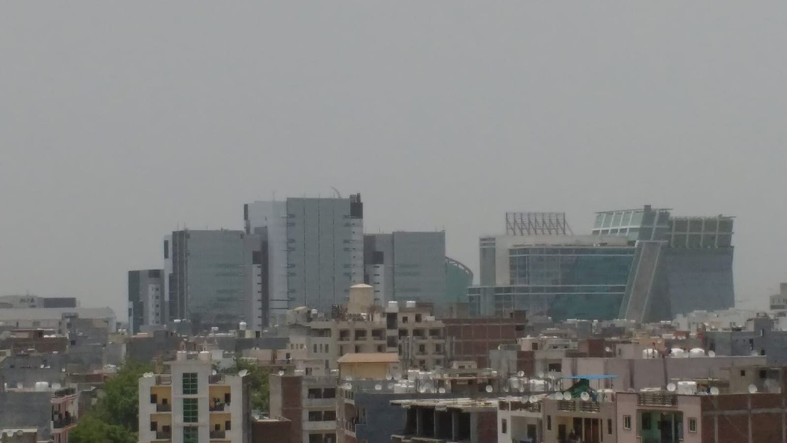 Glass office towers of DLF Cyber City in Gurgaon rise behind low-rise apartment rooftops under a hazy grey sky