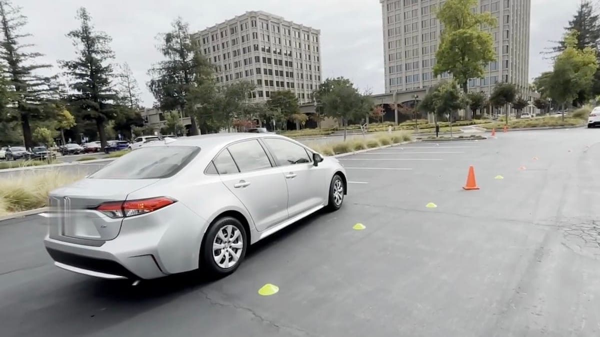 A silver Toyota Corolla driving through an outdoor cone course in a parking lot, with office buildings in the background