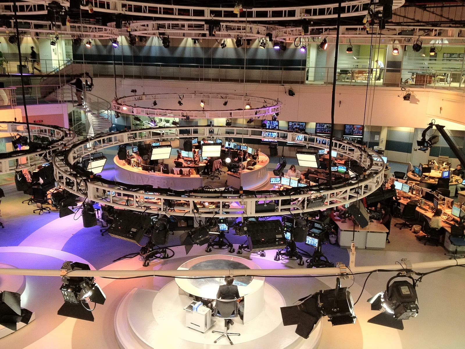 The Al Jazeera English newsroom in Doha seen from above, with a round anchor desk, rows of monitors and studio lights on circular rigs