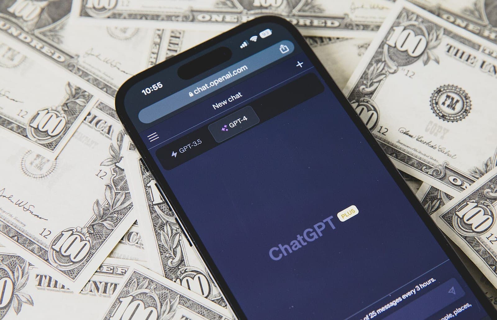 A smartphone showing the ChatGPT Plus web app, lying on a pile of 100 dollar bills.