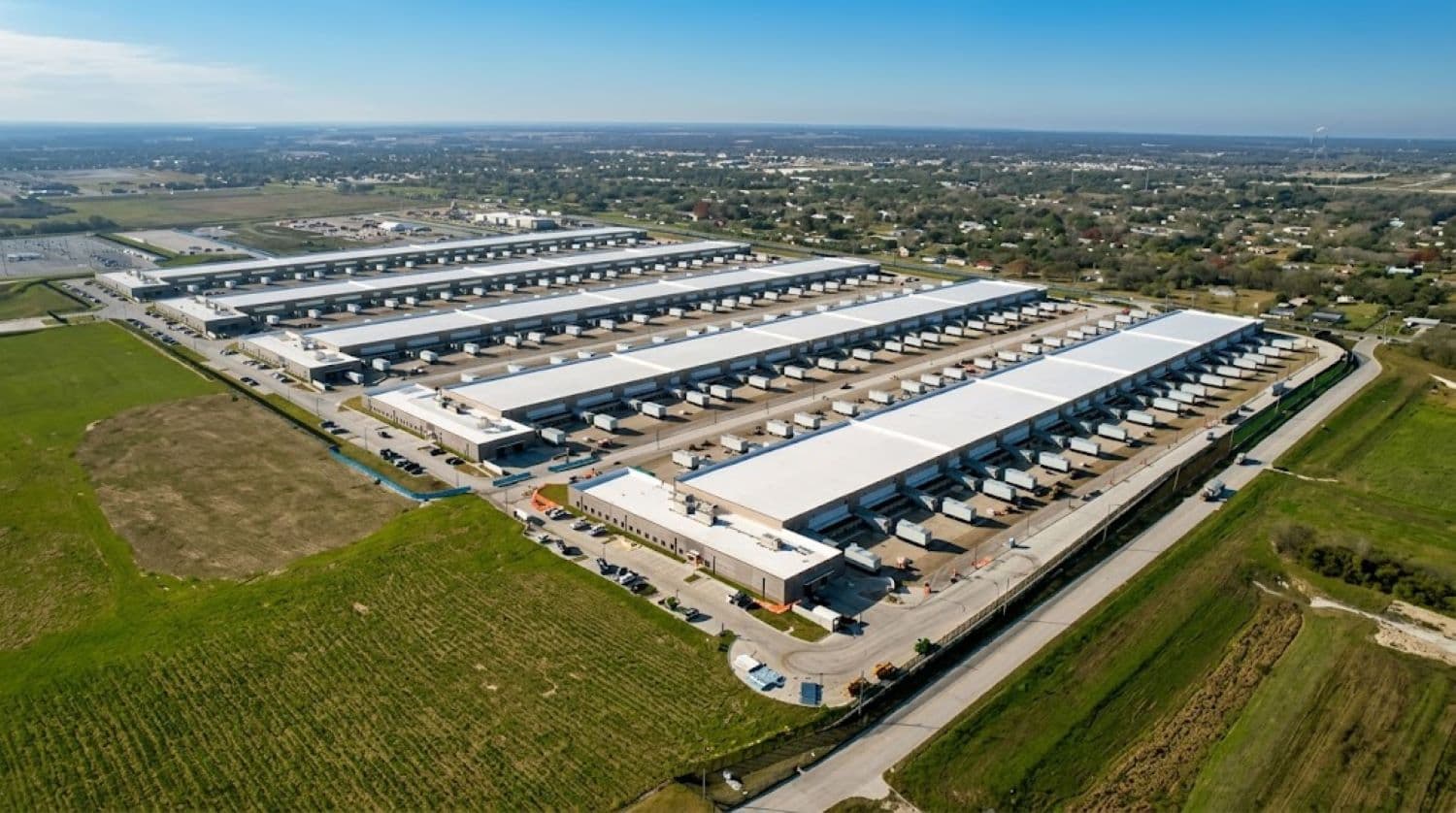 An aerial view of a massive white-roofed industrial data center complex with multiple long warehouse-style buildings, surrounded by open fields and a nearby residential area.