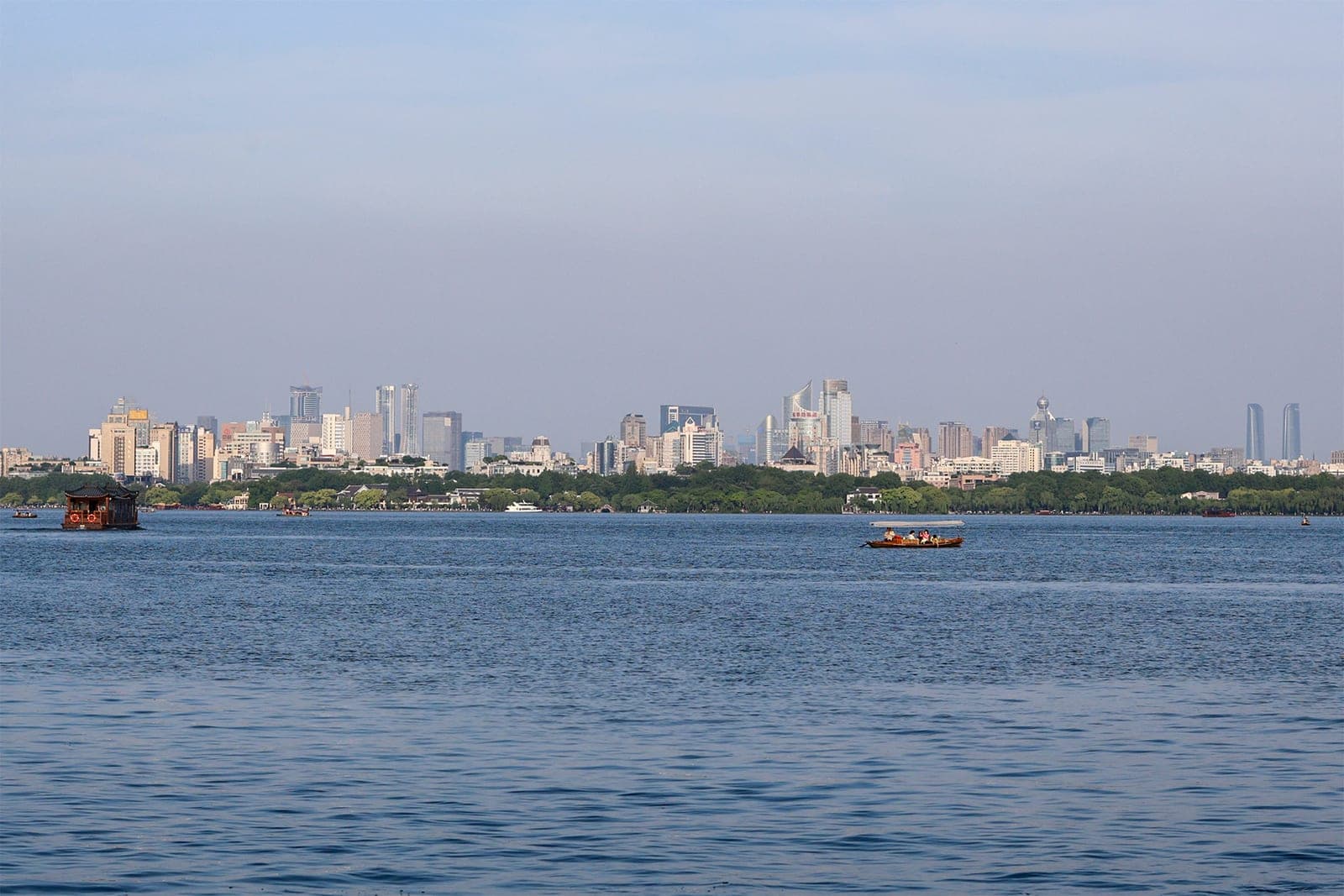 The Hangzhou city skyline seen across West Lake, with small tourist boats on the blue water in the foreground