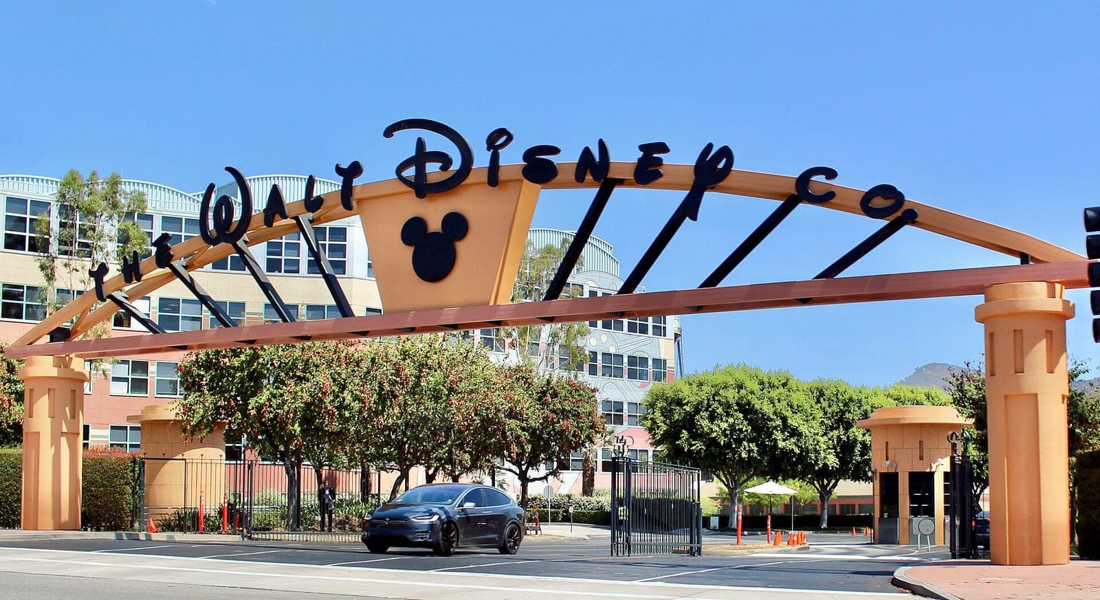 The arched Walt Disney Co. sign over the entrance gate of the Walt Disney Studios in Burbank, with office buildings and trees behind it and a car passing below